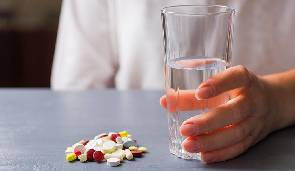 Hand holding glass of water near colorful different capsules and pills on table. Patient taking medicines.